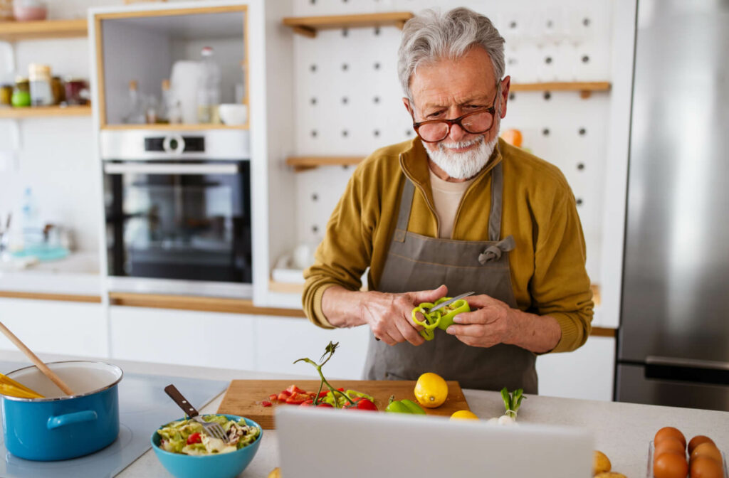 A senior man slicing vegetables in his kitchen while reading a recipe from his laptop.
