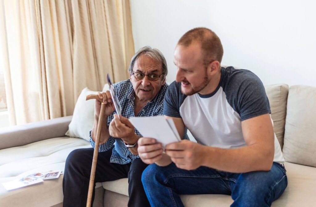 An older adult leans on their cane and nudges their adult child while laughing and looking at photos together
