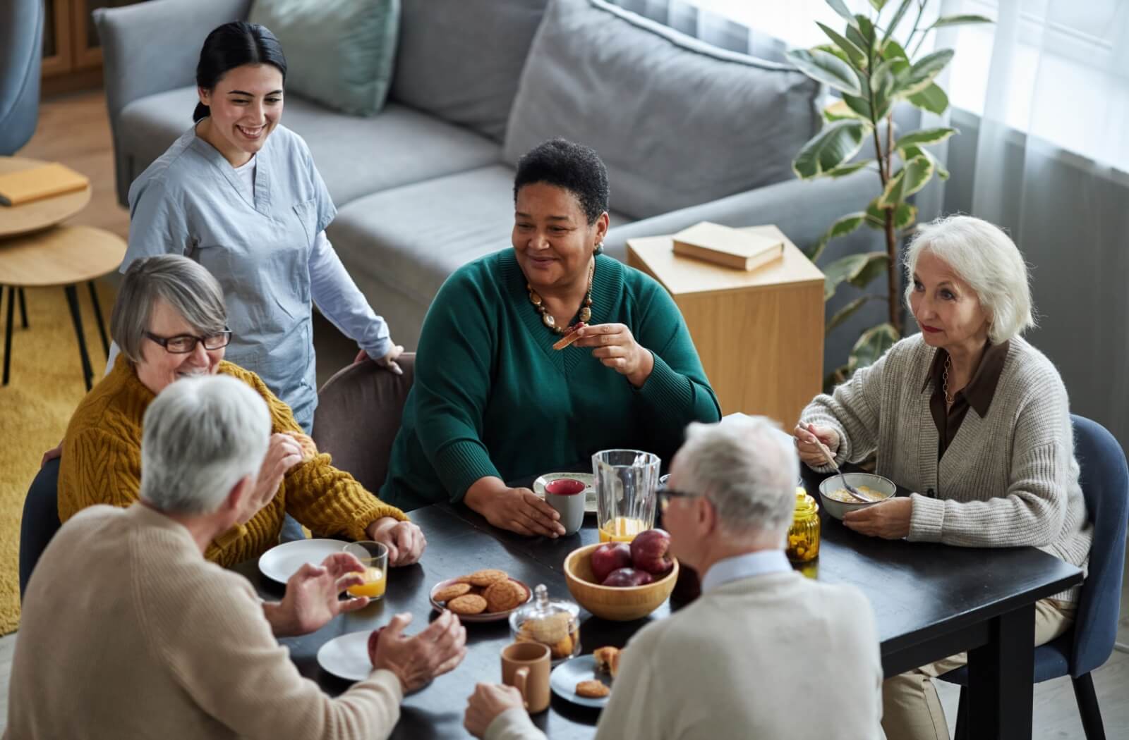 A group of older adults sitting around a breakfast table and talking happily with a nurse standing next to the table, smiling