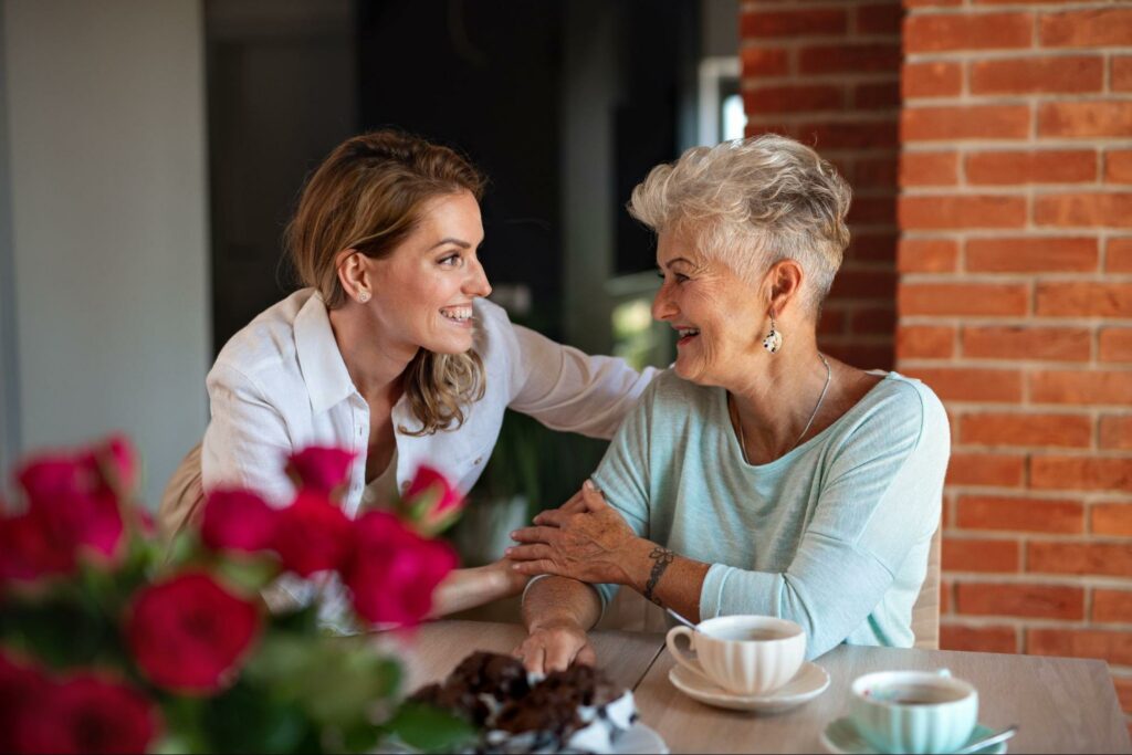 A senior parent and adult daughter sit at a table together, drinking coffee and discussing their plans for an upcoming move. The table is on an outdoor patio, with a potted plant in the center of the table.