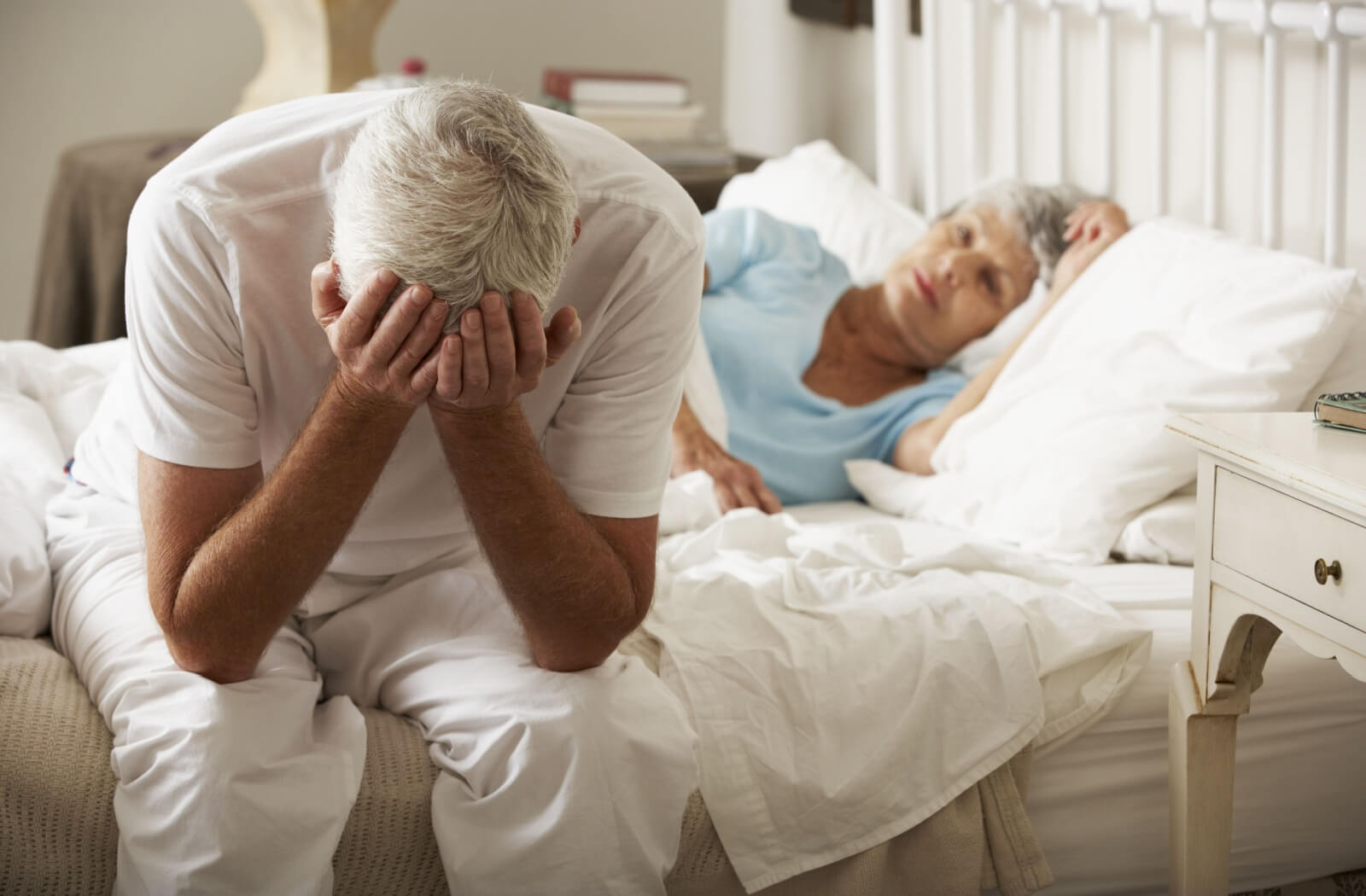 A senior couple in bed, one lying under the covers while the other sits on the edge, head in their hands in embarrassment