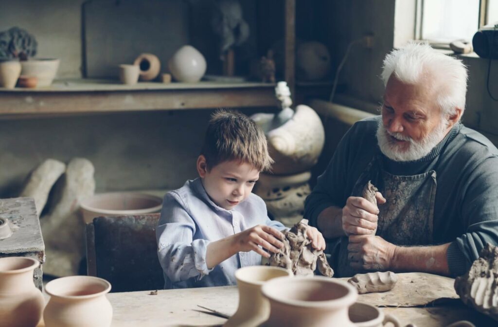 An older adult teaches their grandchild how to work with clay to make pots in a garage studio