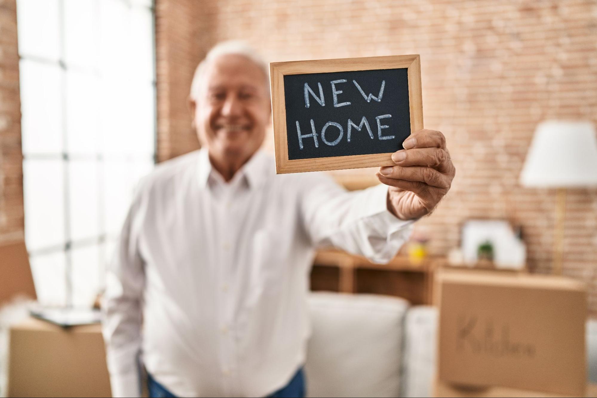 A senior standing in a living room, holding a small chalkboard that has "NEW HOME" written in block letters. The couch behind the senior is stacked with boxes, each labelled with the name of a different room.