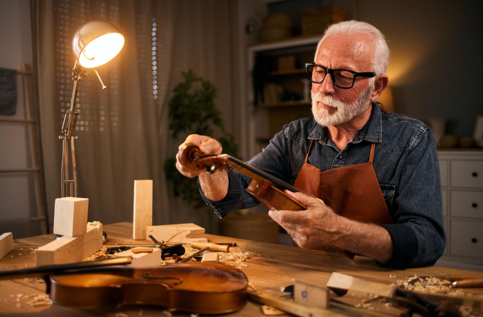 An older individual in black glasses finishes carving the tip of a violin out of wood at an indoor woodshop table