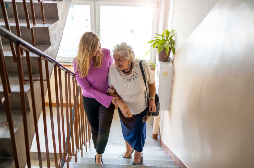 a younger woman holds a senior woman's arm in support while they go upstairs