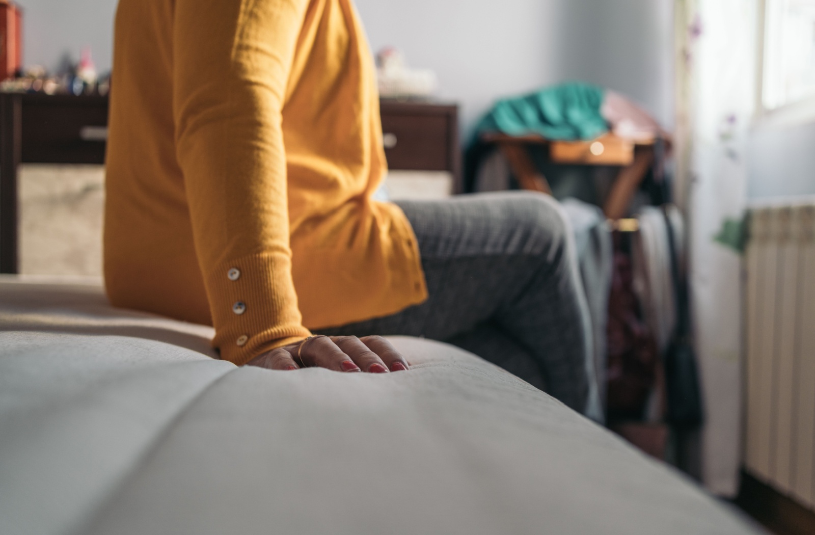 Side view of unidentified older woman (visible from neck down) in yellow cardigan sitting on bed with legs crossed.