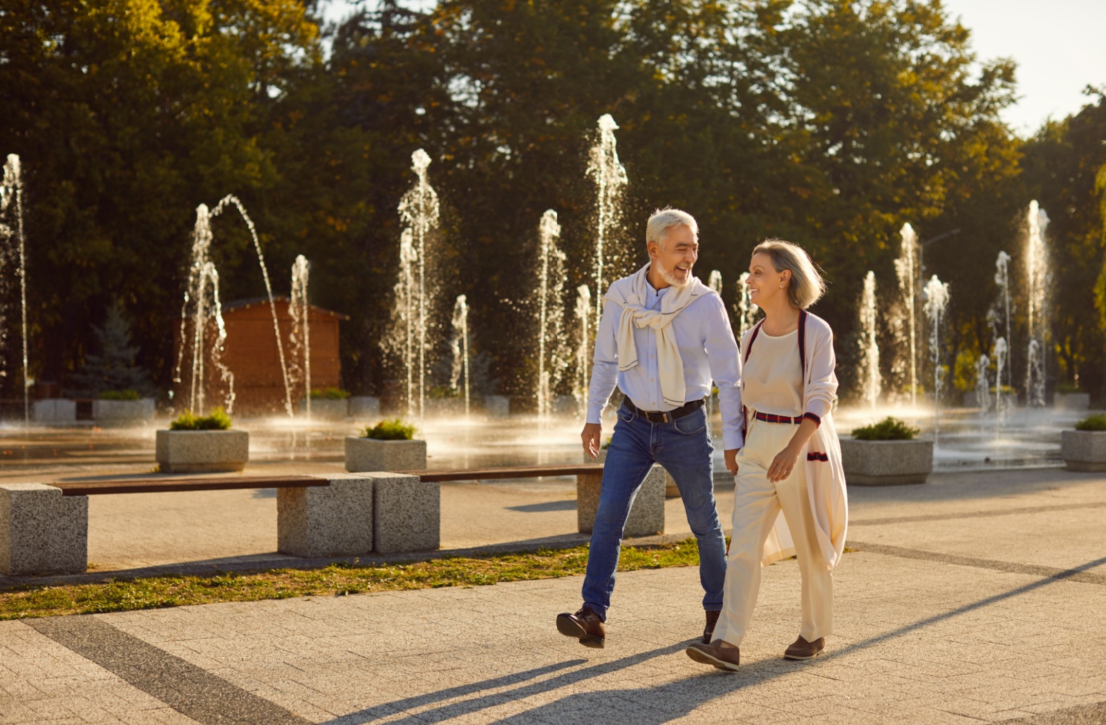 An older couple laughs while walking hand-in-hand through a park full of fountains in the evening