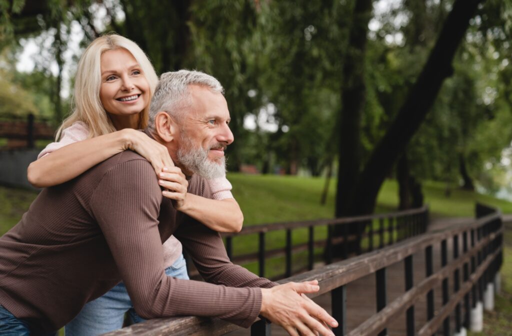 An older adult leans against a bridge's railing and takes a moment to enjoy a green park's scenery while their smiling spouse hugs them from behind