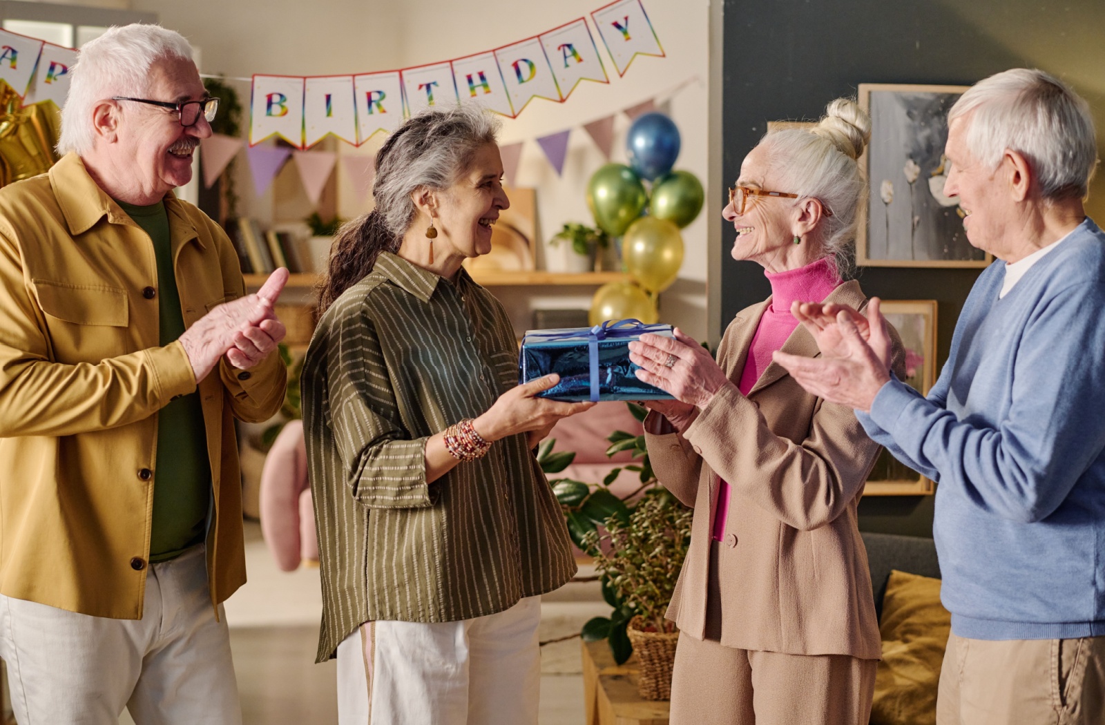 Group of seniors smiling and exchanging a gift during a festive birthday celebration with balloons and decorations